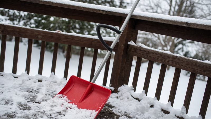 A red snow shovel with a second curved handle leaning on a snowy deck railing, in Portland, Maine